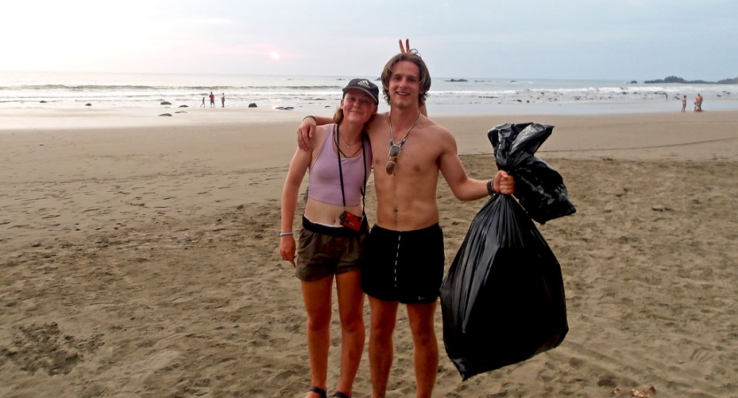 two people stand on a beach after a service project. one of them is holding a trash bag while the other gives them bunny ears.